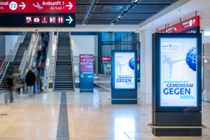 Kampagne: "Gemeinsam gegen Krebs" an einem Flughafen auf digitalen Werbeträgern