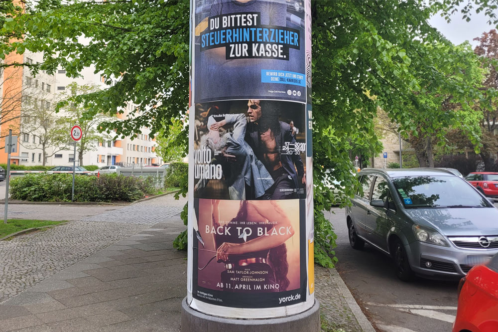 Im Fokus des Bildes steht eine Säule. Auf der Säule ist die Plakatwerbung von Cie. Toula Limnaios zu sehen. A column on the street in Berlin. The column advertisement shows the dance troupe Cie. Toula.