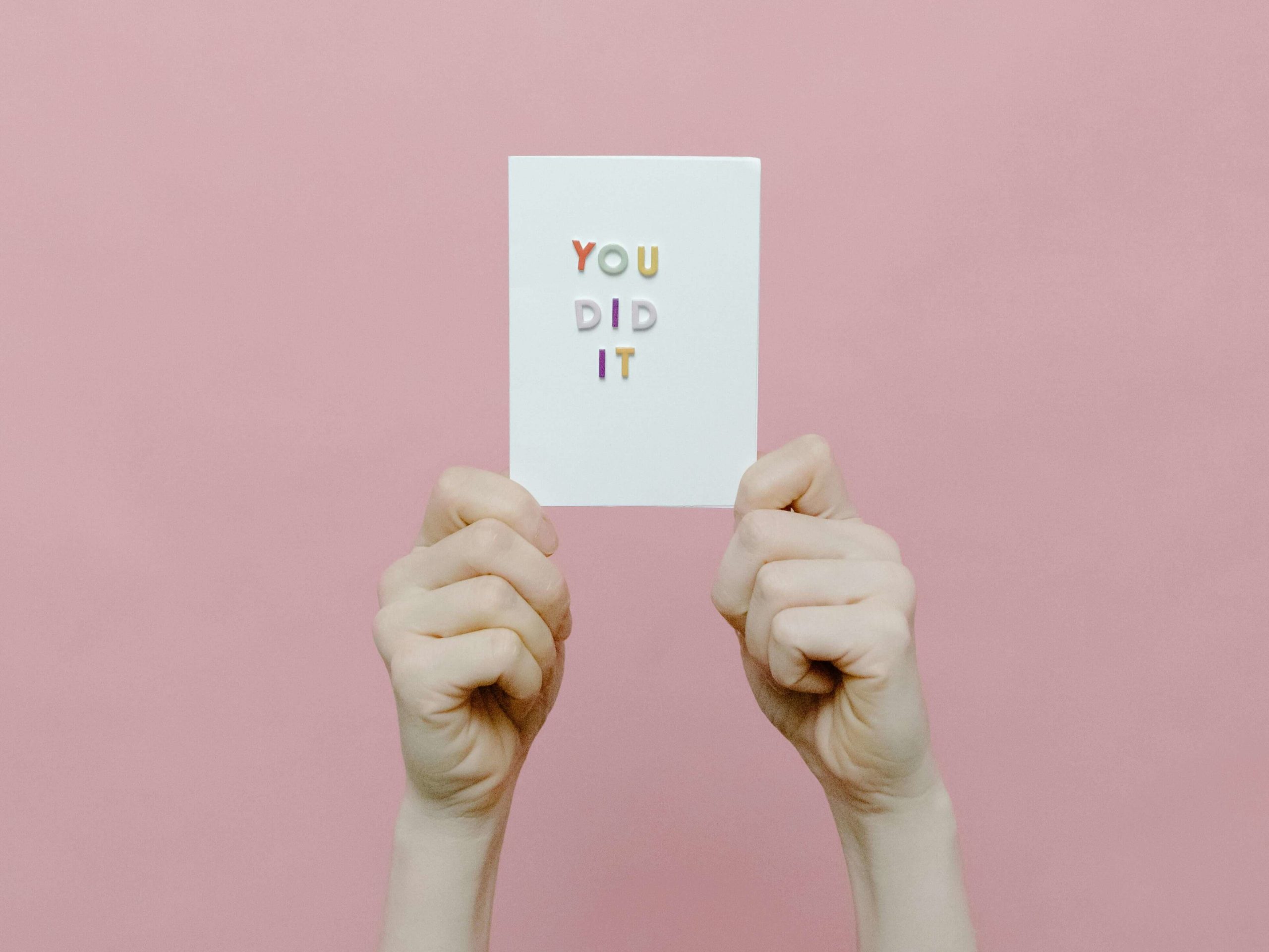 Zwei Hände halten ein Schild mit der Aufschrift „You did it“ vor einen rosafarbenen Hintergrund.