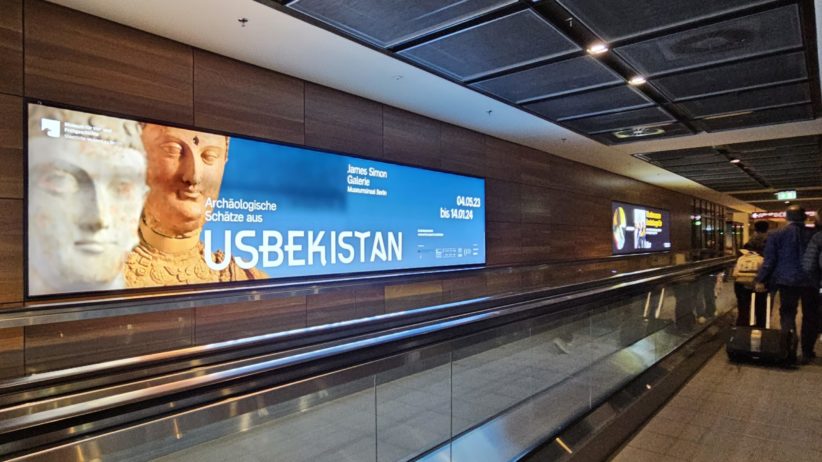 Am Terminal 1 des BER Flughafen Berlin-Brandenburg hängt an der Wand neben einem Laufband ein hinterleuchtetes Coloramen. Die Backlight Werbefläche zeigt die Werbung für eine Ausstellung zu archäologischen Funden aus Usbekistan. Links sieht man Menschen, die mit Ihren Koffern auf dem Laufband laufen.