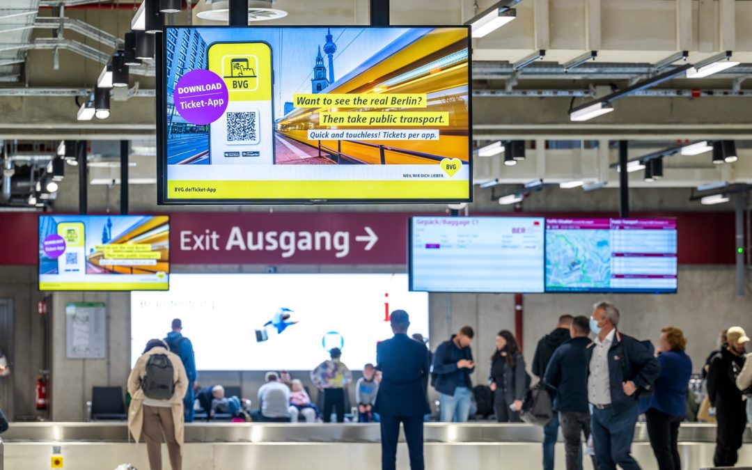 Am Flughafen BER in Berlin hängen über den Terminal 2 Gepäckbändern 2 digitale Screens, die gerade die BVG Ticket App bewerben. Am Gepäckband stehen Menschen und warten auf Ihre Koffer.