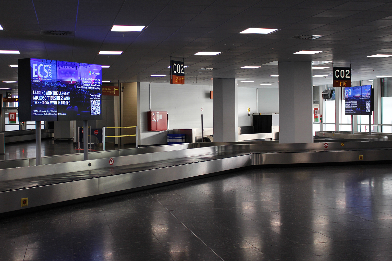 Zwei digitale Bildschirme auf Säulen neben einem Gepäckband am Flughafen. Die Screens werben mit einem lila und blau gestalteten Design und einem QR-Code für den ECS Kongress 2026.