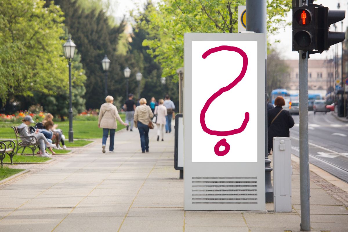 Direkt hinter einem Fußgängerüberweg steht ein freistehendes City Light Poster. Doch anstelle einer Plakatwerbung sieht man nur ein großes rotes Fragezeichen auf weißem Grund. Hinter dem Werbeträger ist eine Bushaltestelle, ein Park mit Wiese, Bäumen und auf Bänken sitzenden Menschen, sowie ein gut besuchter Gehweg und eine Straße.
