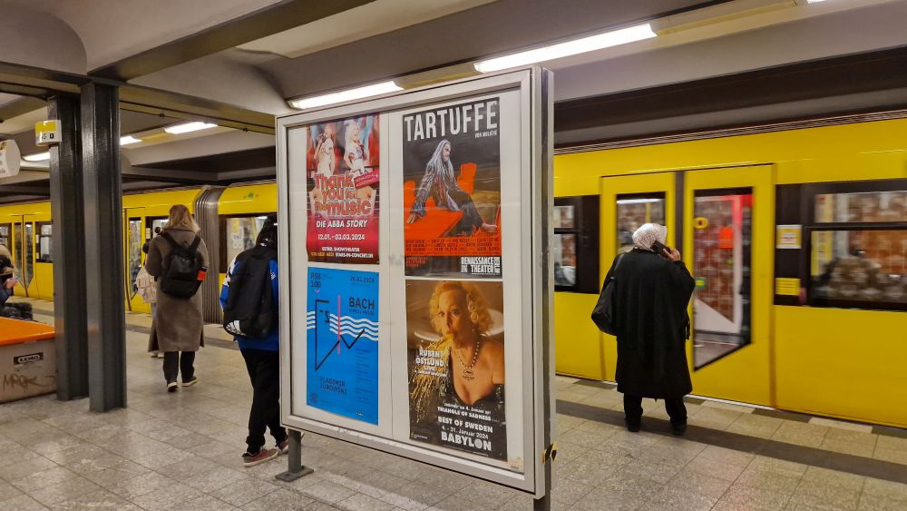 Auf einem U-Bahnhof in Berlin ist eine gelbe U-Bahn eingefahren und Fahrgäste sind zum Einsteigen bereit. Im Vordergrund steht eine Plakatvitrine. In der Allgemeinstelle hängen 4 bunte A1 Plakate, die für Veranstaltungen werben.