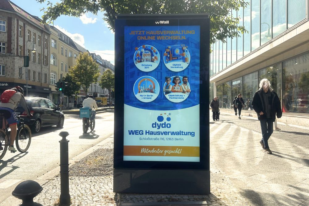 Im Bild sieht man zentral ein digitales City Light Poster. Auf der linken Seite ist die Straße, wo Fahrradfahrer und Autos vorbeifahren. Links laufen Passanten vorbei. Die digitale Werbung zeigt die Werbung von Dydo. The picture shows a digital city light poster in the center. On the left is the street where cyclists and cars pass by. Pedestrians walk past on the left. The digital advertisement shows the Dydo advertisement.
