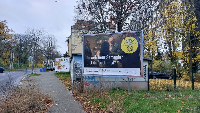 Im Mittelpunkt ist eine Großfläche zu sehen auf eine Wiese in Magdeburg. Ein Fußgängerweg und die Straße sind links zu erkennen. Auf der Plakatwerbung ist zu erkennen die fbb,, die für ein Beratungsnetzwerk für Studierende die abbrechen wollen, helfen.