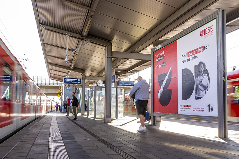 Grossflaechen Werbung - Eine Grossfläche auf ein Münchner Bahnhof. Links des Bildes ist die Bahn und am Bahnhof stehen Menschen. Es zeigt eine Werbung über die IESE Business Schule.