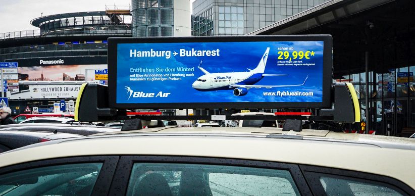 Taxiwerbung Berlin : Digitaler Screen auf einem Taxi Dach. Querformat. Die Taxiwerbung zeigt ein Flugzeug und wirbt für ein Flugangebot von Hamburg nach Bukarest. Weiße und gelbe Schrift auf blauem Hintergrund.