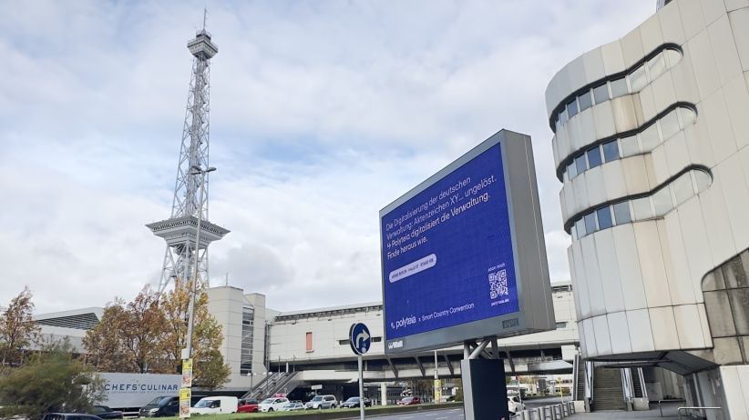 wtm-aussenwerbung- Ein digitales City Light Board steht rechts neben einer Straße in Berlin. Das Werbemotiv ist blau und von der Firma Polyteia. Links im Hintergrund ist der Funkturm von der Messe Berlin zu sehen.