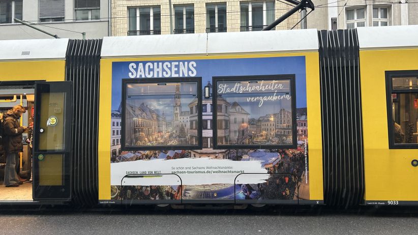 wtm-aussenwerbung tram Berlin - Eine gelbe Tram in Berlin hat auf der Seite ein Traffic Board. Das Werbemotiv zeigt ein Weihnachtsmarkt. Im Hintergrund sind Gebäude zu erkennen.