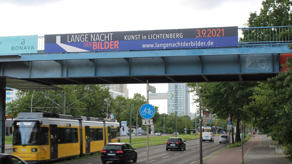 An einer Brücke in Lichtenberg hängt Brückenwerbung, auf der für die Lange Nacht der Bilder geworben wird. Unter der Brücke fahren Autos und eine Tram drunter durch.
